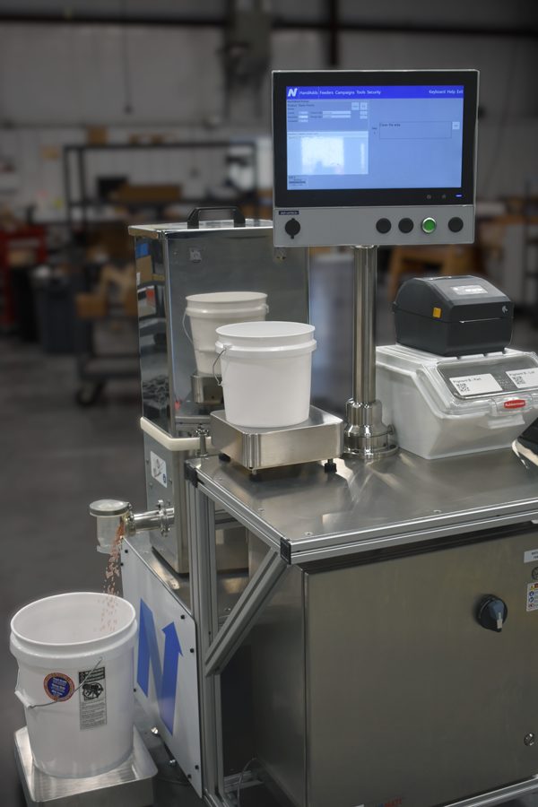 Stand BatchMATE con integración del alimentador. Muestra la estación automatizada de dosificación de microingredientes completa con pantalla, dos básculas, un alimentador, escáner e impresora.